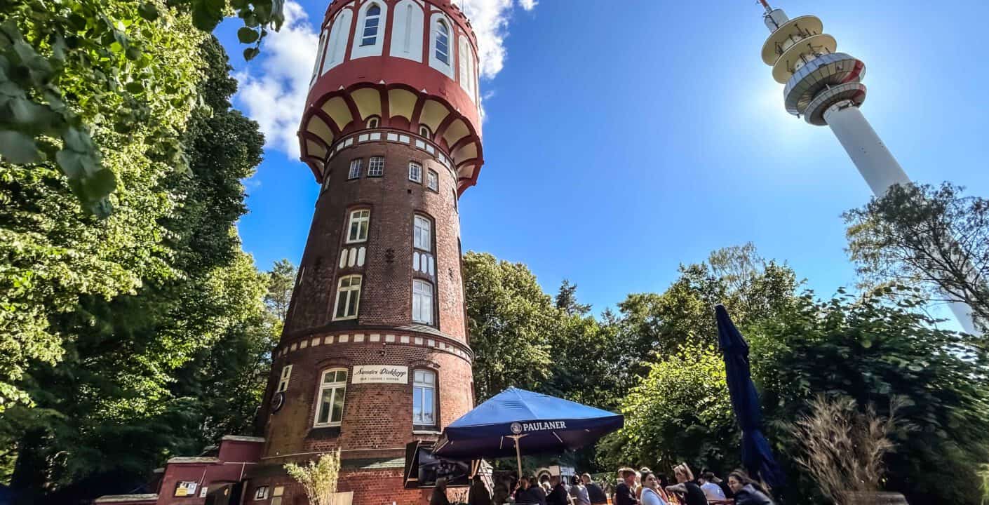 Alter: Historischer Wasserturm bei Sonnenschein mit Außenbestuhlung und Gästen, umgeben von grünen Bäumen, in Düsseldorf, Deutschland, bei gutem Wetter.