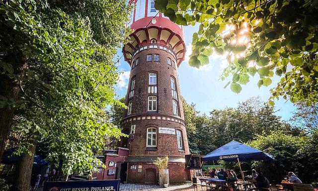 Alt Text: Historischer Wasserturm in grüner Parkumgebung mit Sonnenlicht, darunter ein Biergarten mit Gästen und Sonnenschirmen, im stilvollen Foto.