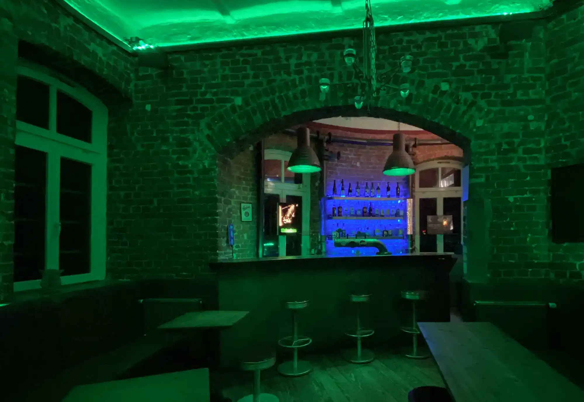Kellerbar mit grüner und blauer Beleuchtung, offener Thekenbereich, rustikale Backsteinwände, gemütliche Sitzgelegenheiten, ideal für Partys und Treffen, modernes und angesagtes Ambiente, ideal für private Feiern oder Events.