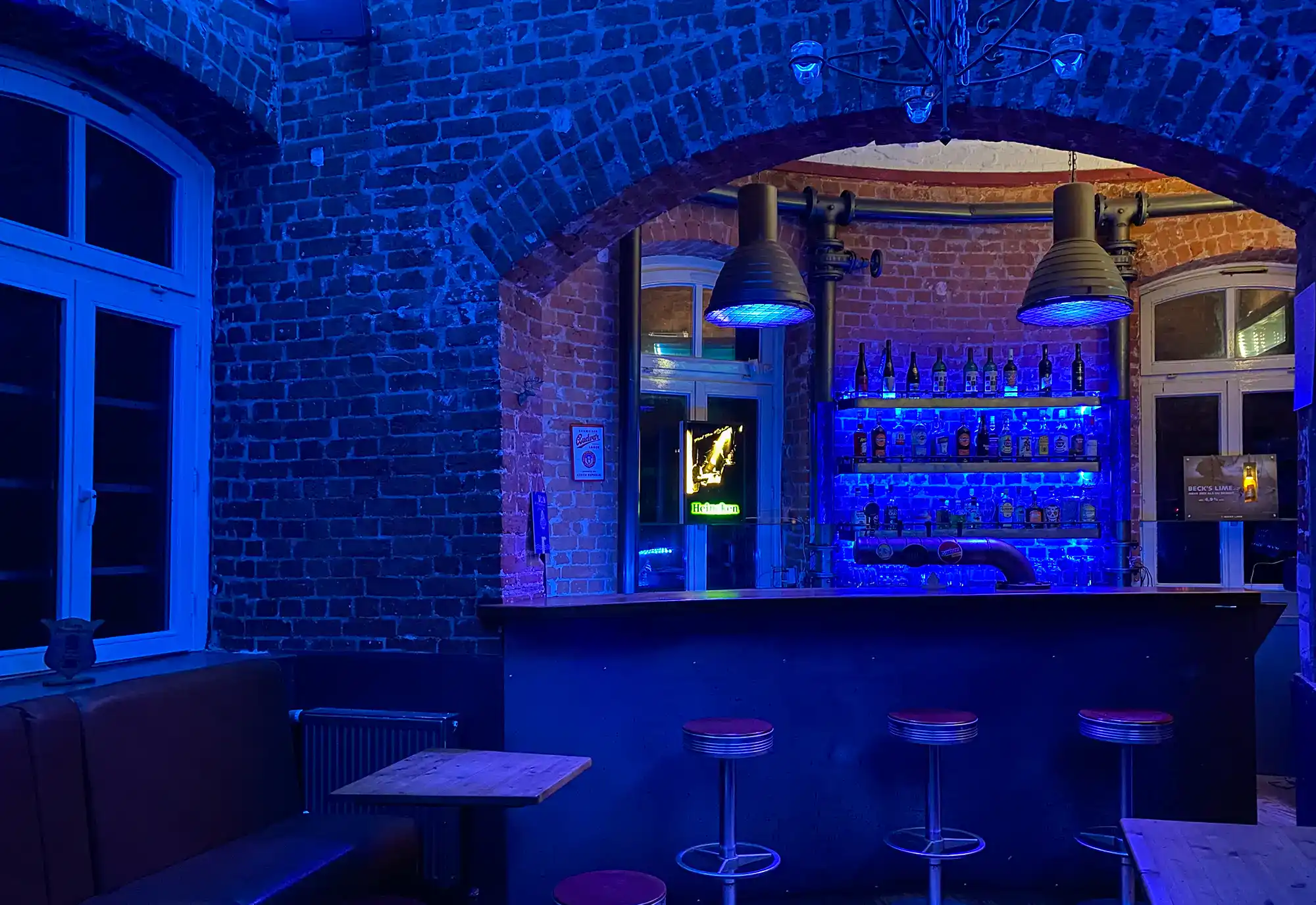 Moderne Bar im Industrie-Design mit Backsteinwänden, blauer LED-Beleuchtung, Steel-Armaturen und stylischen Barhockern, ideal für stilvolle Abende und Drinks in Berlin.