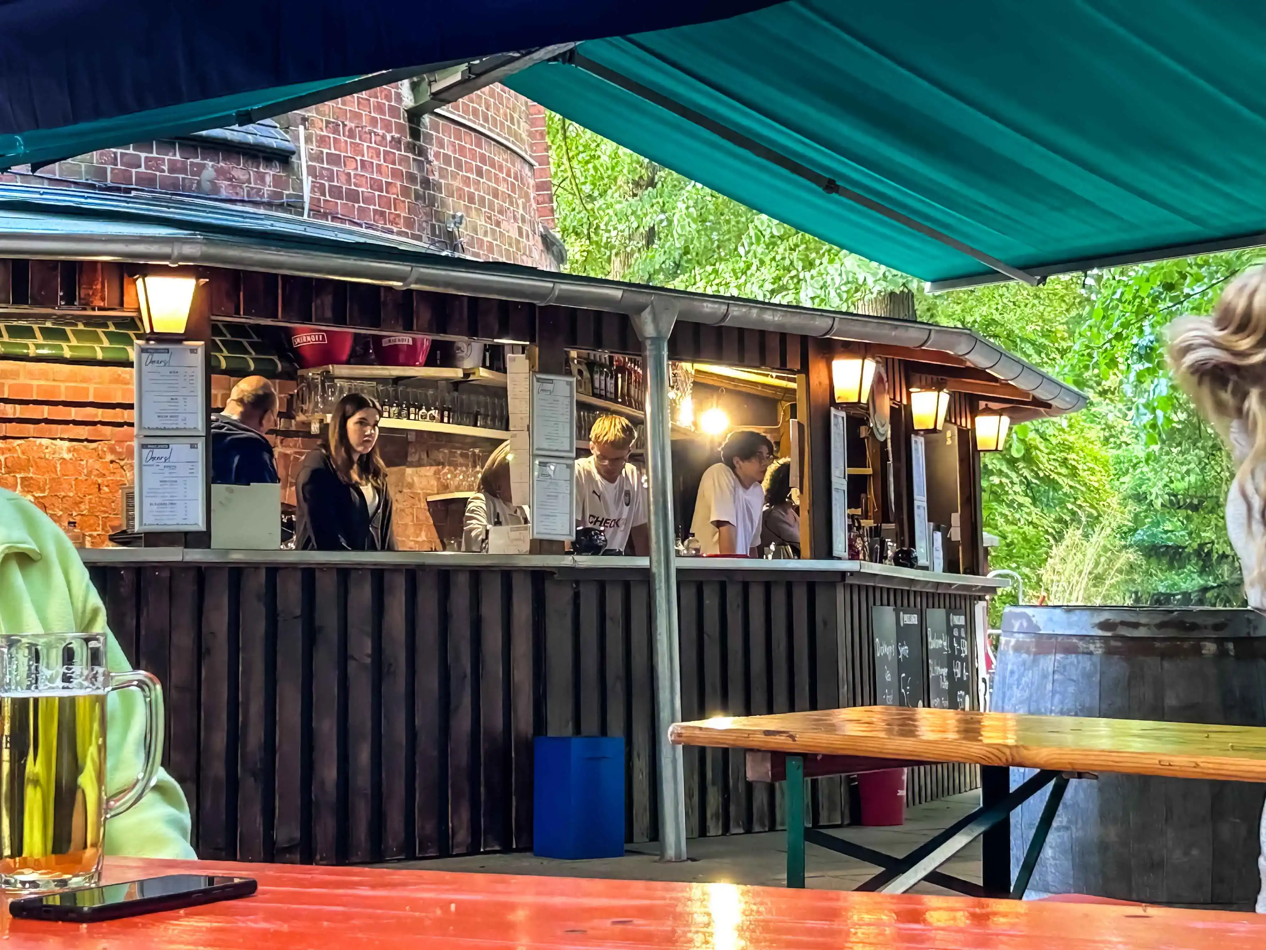 sander dickopp 12 Gemütliche Außengastronomie mit Bar, grünen Bäumen im Hintergrund, Menschen beim Bestellen, Getränk auf dem Tisch, sonnige Atmosphäre, entspannte Stimmung, Entwicklung eines modernen Cafés.