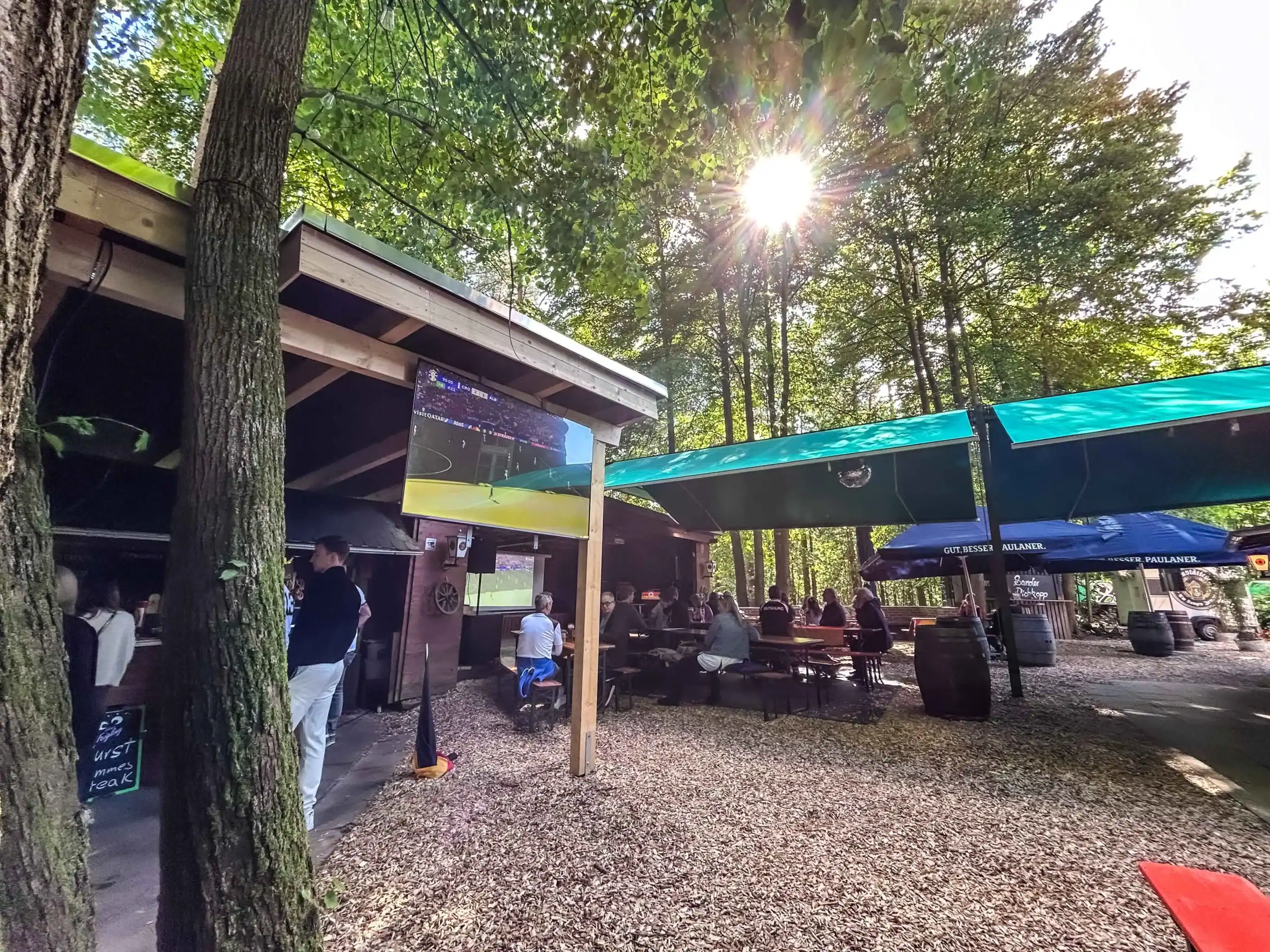 sander dickopp 16 Stimmung im rustikalen Biergarten unter Bäumen, draußen sitzen Menschen beim Public Viewing eines Fußballspiels, entspannte Atmosphäre, Sonnenlicht durch Blätter, sommerliches Freizeit-Event in der Natur.